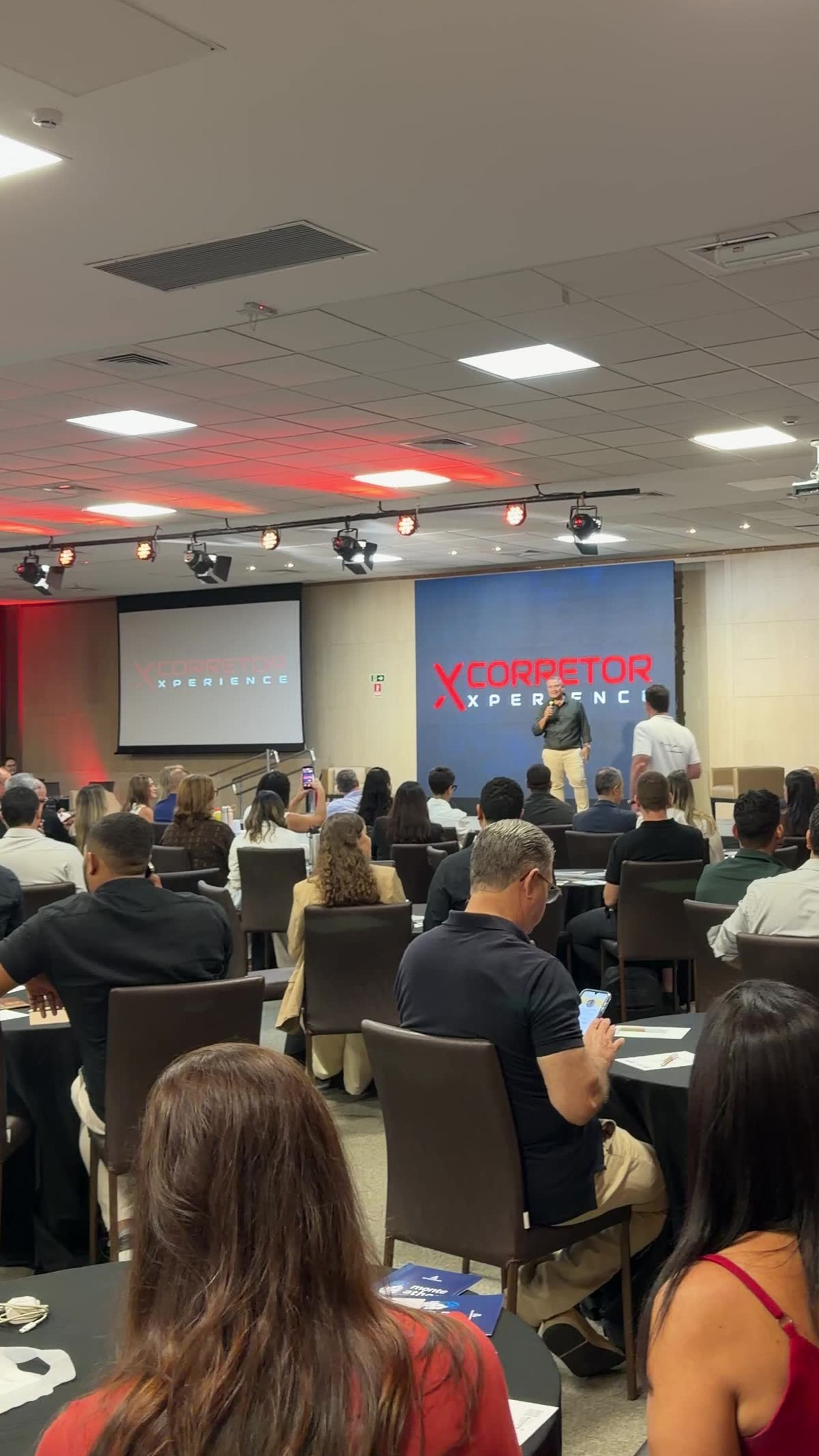 Salão do evento visto de trás da plateia: palco ao centro e telas laterais exibindo a marca Corretor Xperience.